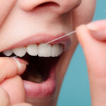 Closeup of woman using string floss to clean between her teeth against a blue background