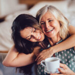 A woman embraces her aging mother from behind as she holds a white mug and smiles in jonesboro