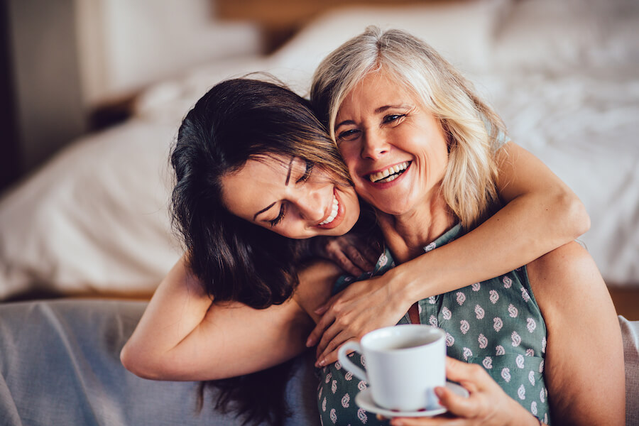 A woman embraces her aging mother from behind as she holds a white mug and smiles in jonesboro